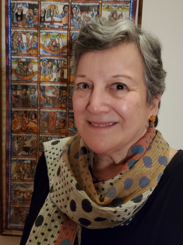 Kay Shelemey in a multicolored scarf, standing near a tapestry