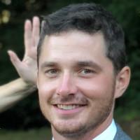 headshot of Henry Pusa standing outdoors, with a hand in the background