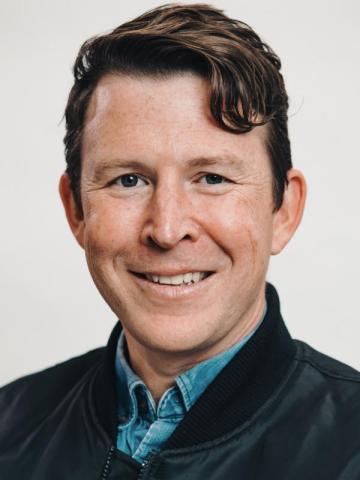 Nick Kawa in a black jacket and blue shirt, with a white background