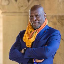 Ato Quayson standing with his arms crossed with an orange scarf and blue jacket