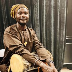 Mohamed Alidu wearing brown, holding a drum, near a curtain