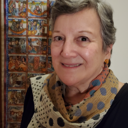 Kay Shelemey in a multicolored scarf, standing near a tapestry