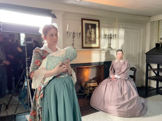 two women in victorian dresses with a filming light behind them and technicians in the shadows