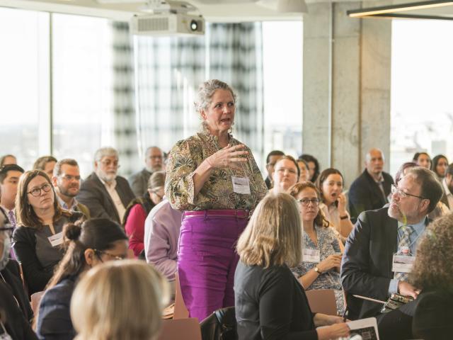 woman standing, speaking in a crowd that is sitting and looking at her
