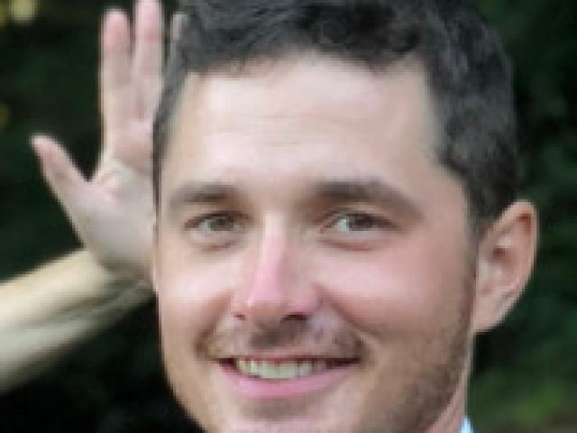 headshot of Henry Pusa standing outdoors, with a hand in the background